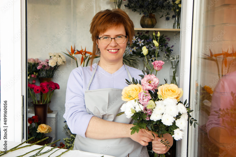 Startup, small business, flower shop. Female florist in apron holds bouquet of flowers for client, at front door of plants studio. 