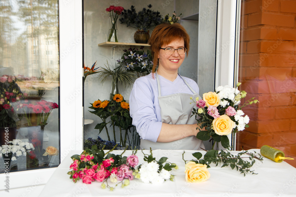 Startup, small business, flower shop. Female in apron holds bouquet of flowers for client and making a bouquet at front door of plants studio. Close up of beautiful flowers lying on table