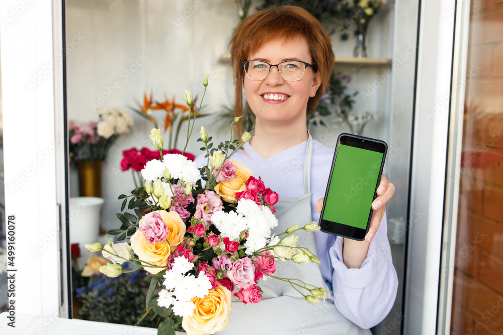 Startup, small business, flower shop. Female florist in apron holds bouquet of flowers for client and mobile phone with green screen, at front door of plants studio. Startup, small business, flower shop. Female florist in apron holds bouquet of flowers for client and mobile phone with green screen, at front door of plants studio.