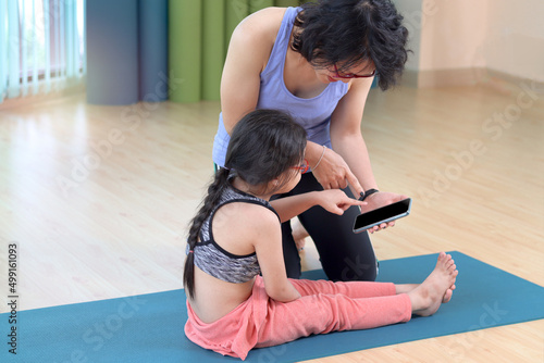Mother and daughter do yoga exercise at indoor studio, use mobile phone for online learning, sporty family study and practice yoga together, mom helping and teaching her child to do yoga pose. photo
