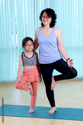 Mother and daughter doing yoga exercise at indoor studio, sporty family practicing yoga together, mom helping and teaching her child to do yoga pose, work out for taking care of body.