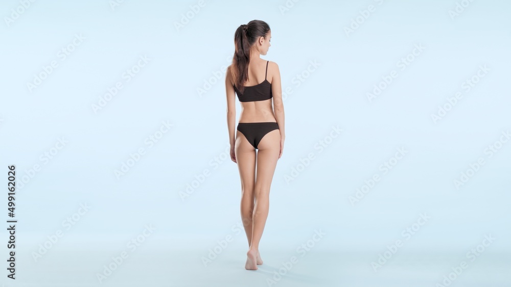 Full shot of young slim dark-haired woman with long ponytail in black underwear stands on her tiptoes back to the camera on pale blue background | Leg skin care concept