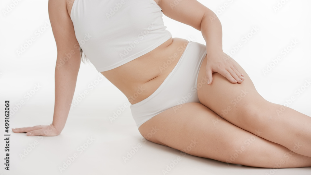 Medium long shot of white-skinned plus size woman in white underwear strokes her leg sitting on the floor on white background | Smooth skin, body and leg care concept