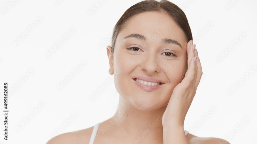 Close-up beauty portrait of young Caucasian woman strokes her cheek and temple smiling wide for the camera on white background | Face care concept Close-up beauty portrait of young Caucasian woman strokes her cheek and temple smiling wide for the camera on white background | Face care concept