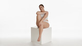 Full-length shot of young Caucasian plus size woman in white underwear strokes her arm sitting on white cube platform and smiling wide for camera on white background | Body care concept