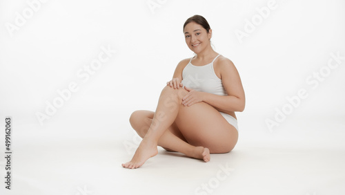 Young Caucasian plus size woman in white underwear strokes her leg skin sitting on the floor on white background | Leg skin care concept