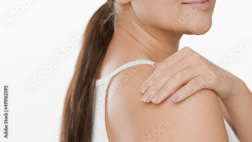 Close-up shot of white-skinned woman with long brown hair in ponytail in white top touches shoulder on white background | Skincare concept
