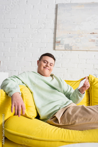 Positive teenager with down syndrome holding smartphone while sitting on couch at home.