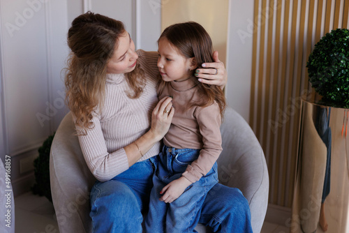 Lovely family mother and daughter embracing and kissing each other indoors. Happy sweet woman with little 6-year-old girl in casual clothers: turtlenecks and jeans sitting on sofa, hugging and smiling © Tatyana