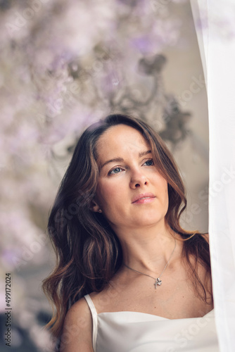 A beautiful young long-haired woman in a light silk dress looks out the window of room decorated with gypsophila trendy color of 2022 year very peri.Selective focus, close up.