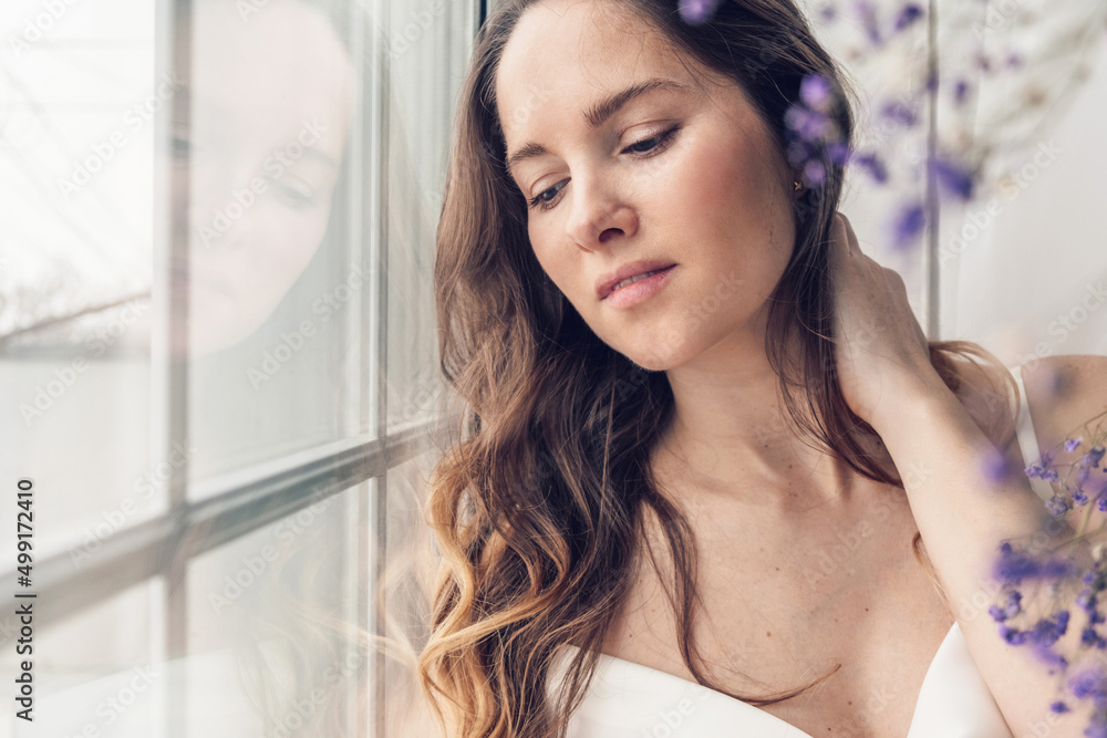 A beautiful young long-haired woman in a light silk dress looks out the window of room decorated with gypsophila trendy color of 2022 year very peri. A beautiful young long-haired woman in a light silk dress looks out the window of room decorated with gypsophila trendy color of 2022 year very peri.