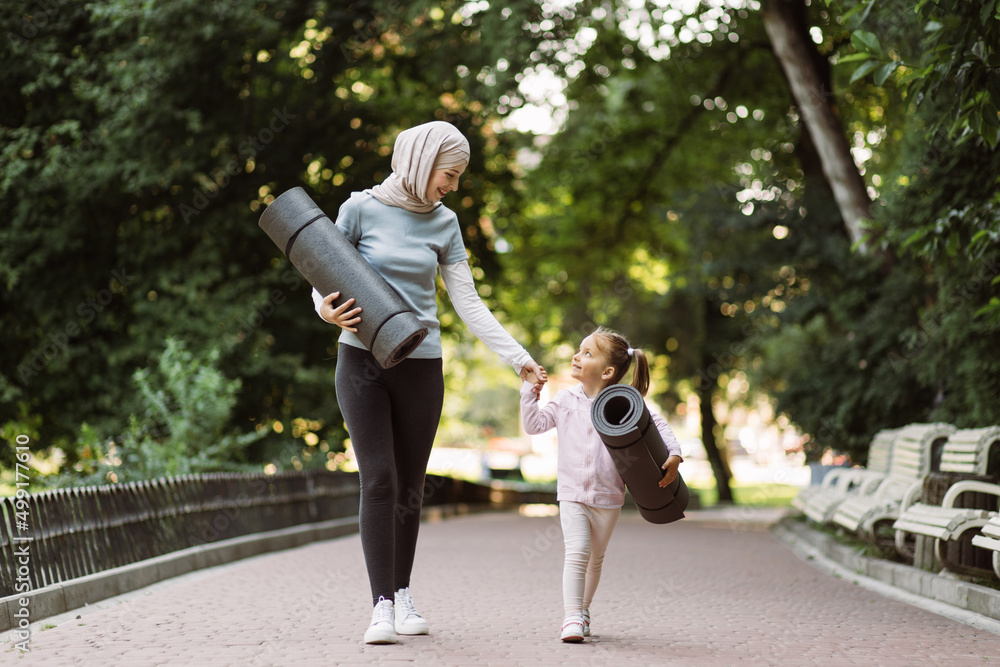 Obraz premium Mother and daughter ready to do exercise outdoors. Likable woman in hijab, walking with her little daughter in the green summer park, holding yoga mats in hands and looking each other