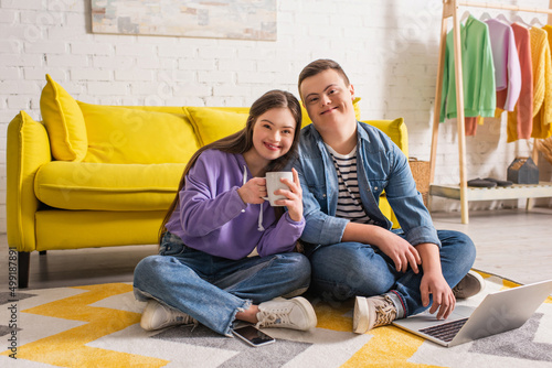 Positive couple of teenagers with down syndrome holding cup and using laptop at home. photo