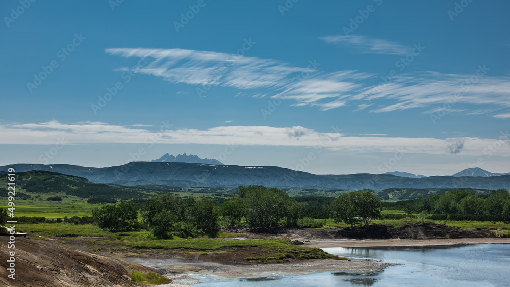 There is a lake in the caldera of an extinct volcano. Green vegetation in the valley. A picturesque mountain range against a background of blue sky and clouds. Kamchatka. Uzon