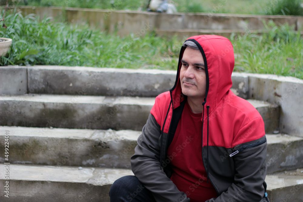 A hooded man sits on concrete steps in a garden.