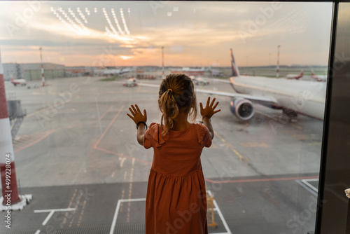 Little girl at the airport is waiting for landing at the big window. Cute kid stands at the window against the backdrop of airplanes. Looking forward to leaving for a family summer vacation