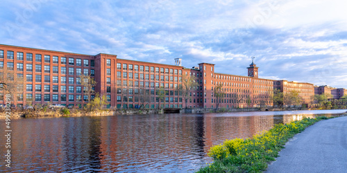 Panorama of a historic cotton mill building with a clock tower in an old industrial park on the Nashua River, illuminated by the sun during sunset in May. Panoramic photography. Nashua, New Hampshire,
