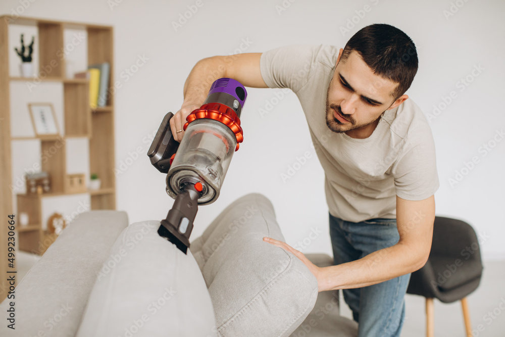 A young man cleans the sofa with a vacuum cleaner