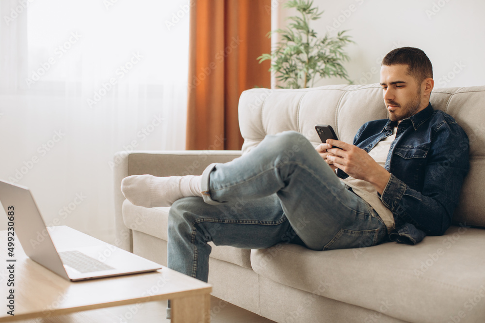 Portrait of smiling guy talking on mobile phone, working on laptop, sitting on the sofa in living room. Portrait of smiling guy talking on mobile phone, working on laptop, sitting on the sofa in living room.
