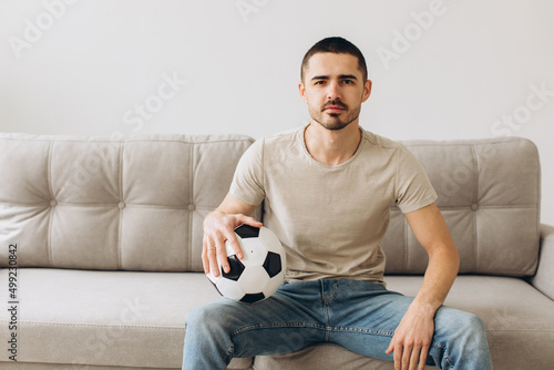 A man sits on a sofa in the living room holding a ball and cheers for the football team and watching a match