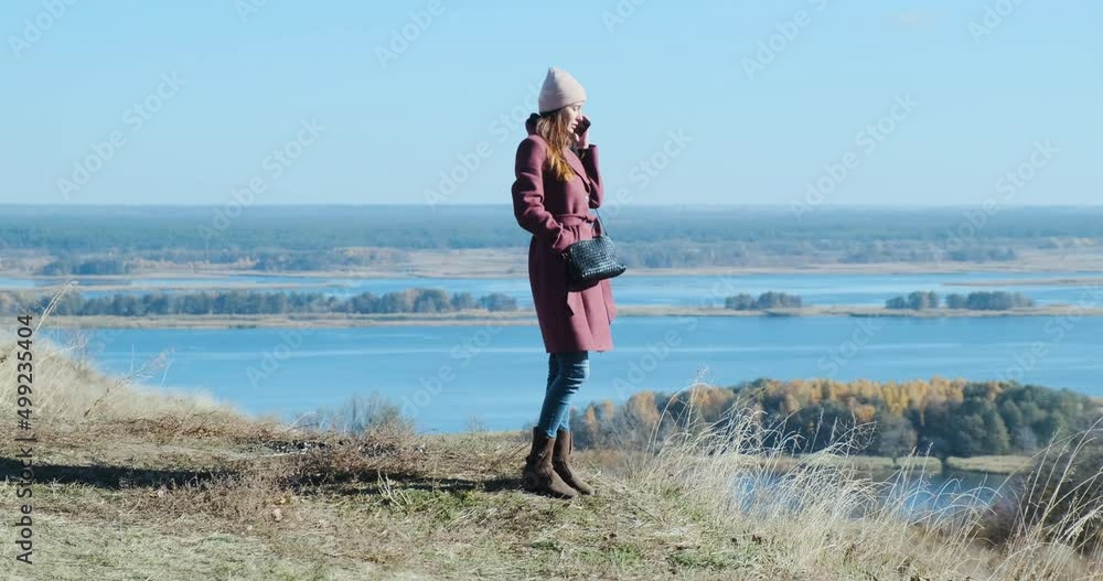 young woman in a coat and hat stands on a hilltop and speaks on the phone in the cold season. Against the backdrop of a wide spreading river with long islands. Medium shot, morning. young woman in a coat and hat stands on a hilltop and speaks on the phone in the cold season. Against the backdrop of a wide spreading river with long islands. Medium shot, morning.