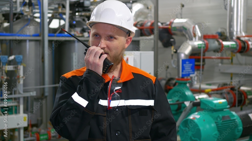 Technological engineer in a white helmet transmits instructions to the staff using a walkie-talkie. A bearded man with blue eyes at a workplace in an industrial workshop. Technological engineer in a white helmet transmits instructions to the staff using a walkie-talkie. A bearded man with blue eyes at a workplace in an industrial workshop.