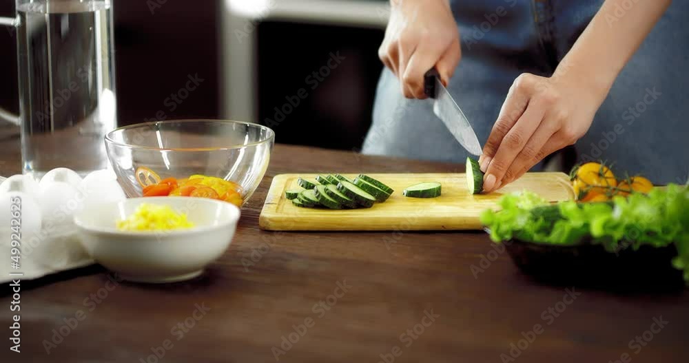 Young woman in jeans is preparing a salad of healthy foods in the kitchen. Women's hands cut a cucumber with a knife on a cutting board. Fresh vegetables on a wooden chopping board. Eco-friendly Young woman in jeans is preparing a salad of healthy foods in the kitchen. Women's hands cut a cucumber with a knife on a cutting board. Fresh vegetables on a wooden chopping board. Eco-friendly
