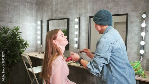 redhaired girl sits and laughs, and makeup artist applies foundation to her face