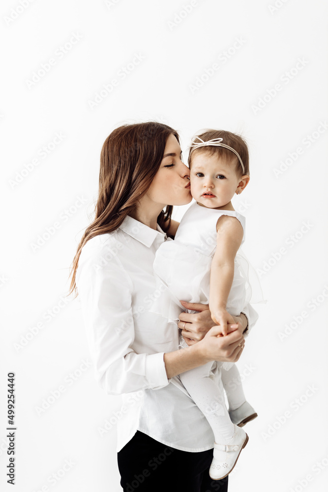 Happy family. Emotional young stylish mother staying and holding in arms laughing adorable toddler baby daughter, having fun playing together, isolated in light room, studio. Happy time of motherhood. Happy family. Emotional young stylish mother staying and holding in arms laughing adorable toddler baby daughter, having fun playing together, isolated in light room, studio. Happy time of motherhood.