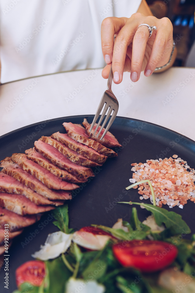 Woman's hand holding fork and eating beef steak with salad, in cafe.