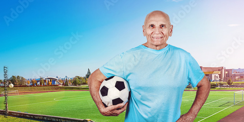 Mature football coach holds black and white ball in hands standing against soccer field with green grass outdoor