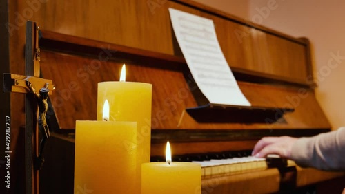 Female musician pianist finishes playing a beautiful religious christian melody on a brown upright piano with musical notes sheet in cozy room or curch with three burning candles and a crucifix and cl photo