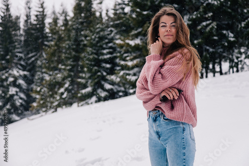 A young, beautiful, long-haired girl in a pink sweater poses in a winter forest in the mountains © Denis