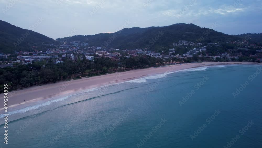 Early dawn on the island of Phuket. Top view from the drone on the sand and waves. People are walking along the beach. Calm environment. Green hills and clouds in sky. Lights of the city are shining Early dawn on the island of Phuket. Top view from the drone on the sand and waves. People are walking along the beach. Calm environment. Green hills and clouds in sky. Lights of the city are shining