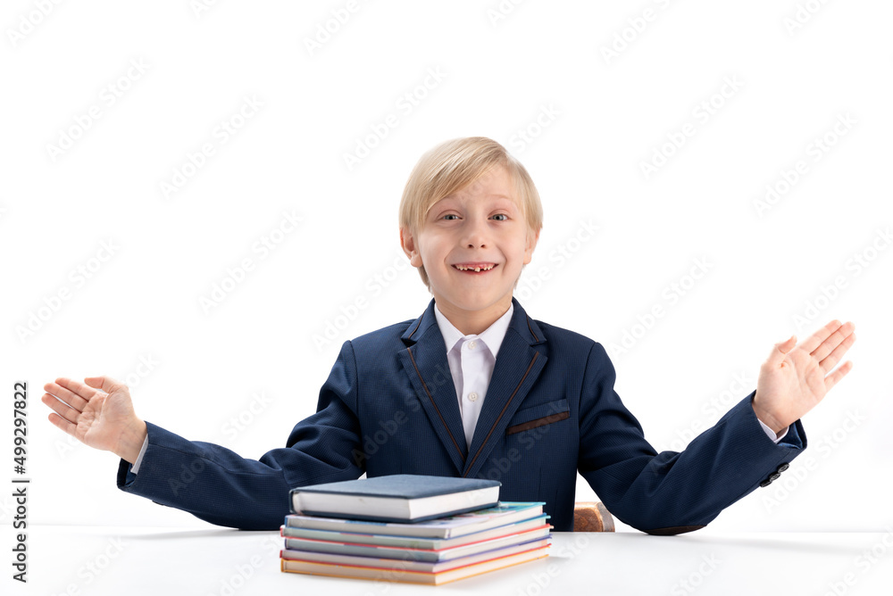 Schoolboy sits at table with stack of notebooks and books and throws up his hands. Boy in school uniform at desk is isolated on white background. Schoolboy sits at table with stack of notebooks and books and throws up his hands. Boy in school uniform at desk is isolated on white background.