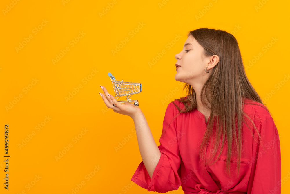 Beautiful young girl in red blouse holds grocery cart in palm of hand on bright orange background. Buyer or customer concept.