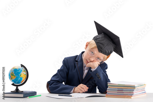 Tired exhausted schoolboy does his homework. Boy in school uniform and student hat sits at table next to notebooks and globe.