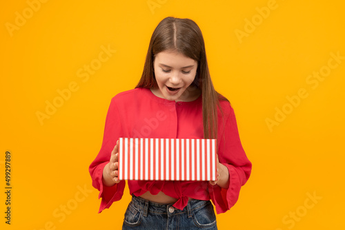 Stunned and surprised young girl looks into an open gift box. Portrait of teenager with gift in hands. Yellow background
