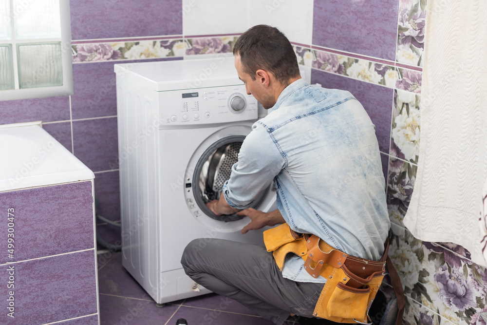 The young handsome repairman in worker suit with the professional tools box is fixing the washing machine in the bathroom The young handsome repairman in worker suit with the professional tools box is fixing the washing machine in the bathroom