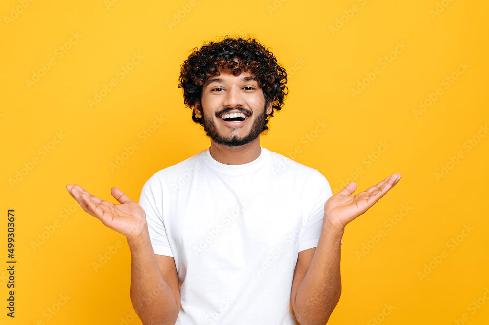 Joyful positive indian or arabian guy with curly hair, wearing white t-shirt, looks at camera happily, spreading his arms to the sides, standing over isolated orange background, looks at camera, smile Joyful positive indian or arabian guy with curly hair, wearing white t-shirt, looks at camera happily, spreading his arms to the sides, standing over isolated orange background, looks at camera, smile