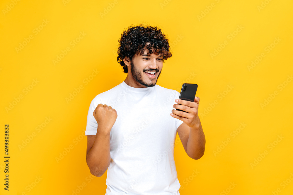 Happy joyful excited Indian or Arabian guy holds smartphone, get unexpected news, winning lottery, stands on isolated orange background, cheerful facial expression, toothy smile, gesturing with fist Happy joyful excited Indian or Arabian guy holds smartphone, get unexpected news, winning lottery, stands on isolated orange background, cheerful facial expression, toothy smile, gesturing with fist