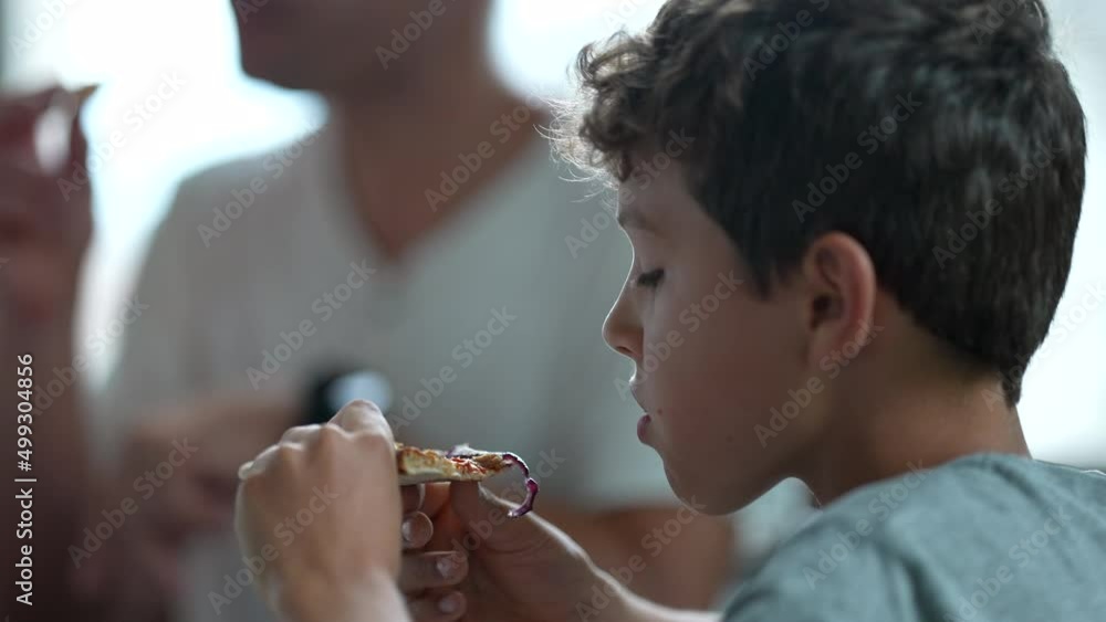 Candid young boy eating piece of pizza with family lunch Candid young boy eating piece of pizza with family lunch