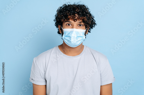 Prevention of infection. Indian or Arabian young adult curly-haired guy, with a protective medical mask on his face, looking at the camera while standing on isolated blue background © Kateryna