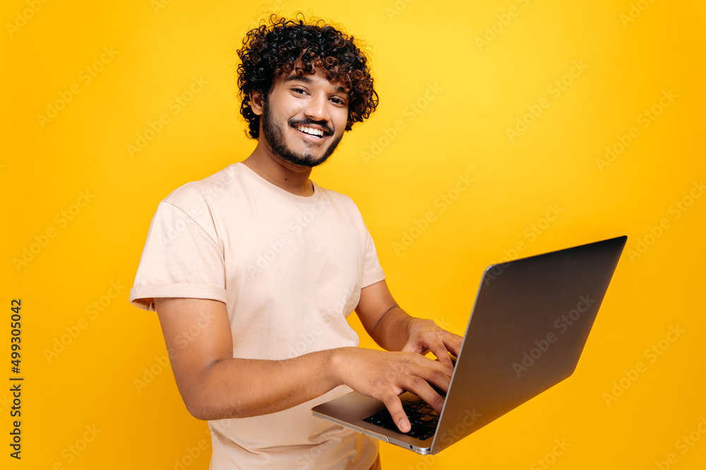 Positive curly-haired indian or arabian millennian guy in t-shirt, typing on laptop keyboard, standing over isolated orange background, looking at camera, smiling friendly. Technology concept Positive curly-haired indian or arabian millennian guy in t-shirt, typing on laptop keyboard, standing over isolated orange background, looking at camera, smiling friendly. Technology concept