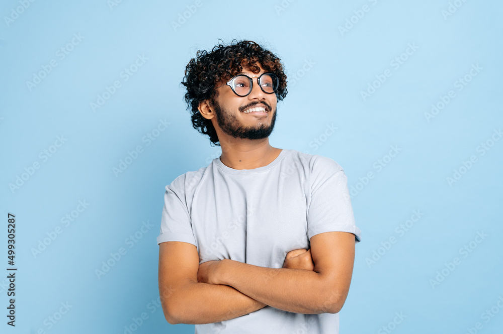 Handsome attractive indian or arabian curly-haired guy, wearing t-shirt and glasses, standing over isolated blue background with crossed arms, looking away, smiling happily Handsome attractive indian or arabian curly-haired guy, wearing t-shirt and glasses, standing over isolated blue background with crossed arms, looking away, smiling happily