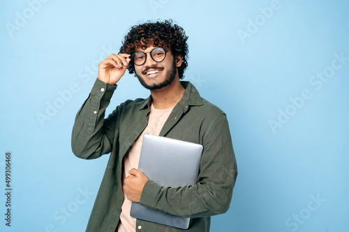 Smart handsome positive indian or arabian millennial guy, with glasses, student or freelancer, holding a laptop in hand, standing on isolated blue background, looking at the camera, smiling friendly