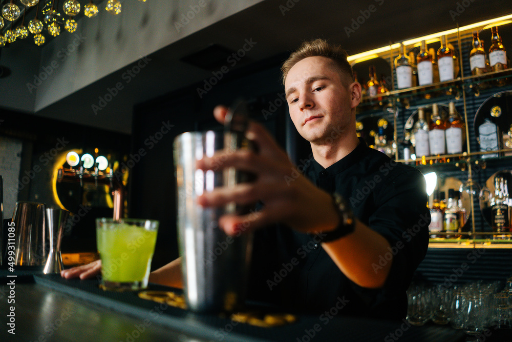 Low-angle view of confident bartender male making colorful alcoholic cocktail standing behind bar counter in modern dark nightclub, on blurred background of shelves with different alcoholic drinks.