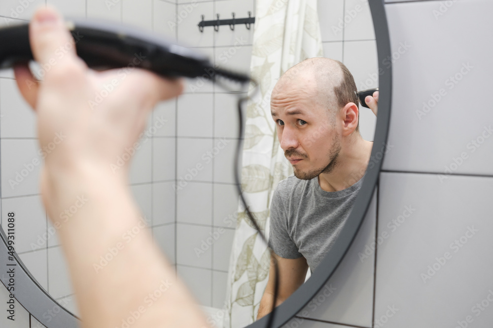 Obraz premium Hair loss problem. Close up of a young caucasian man standing against mirror in the bathroom holding a trimmer and cutting hair by himself.