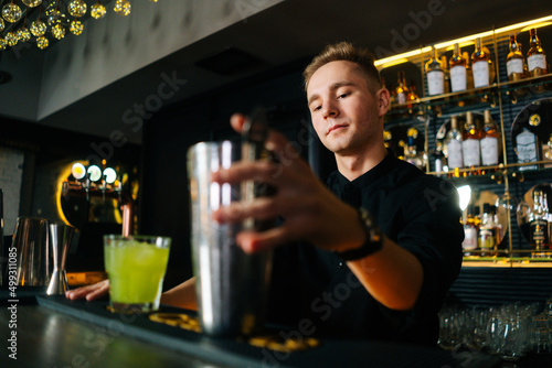 Low-angle view of confident bartender male making colorful alcoholic cocktail standing behind bar counter in modern dark nightclub, on blurred background of shelves with different alcoholic drinks.