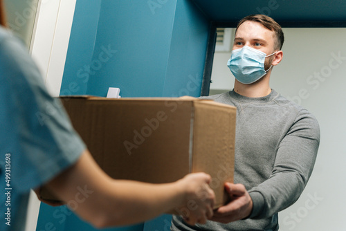 Delivery man wearing medical face mask giving cardboard box parcel to female customer on doorstep at home. Rear view of unrecognizable woman client receiving parcel from courier male at front door